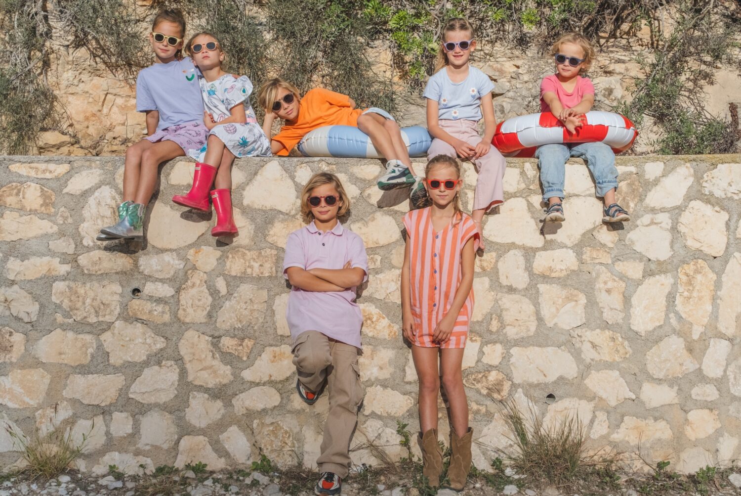 Groep vrolijke kinderen met zonnebrillen OKKY zittend en staand bij een stenen muur in de zon.