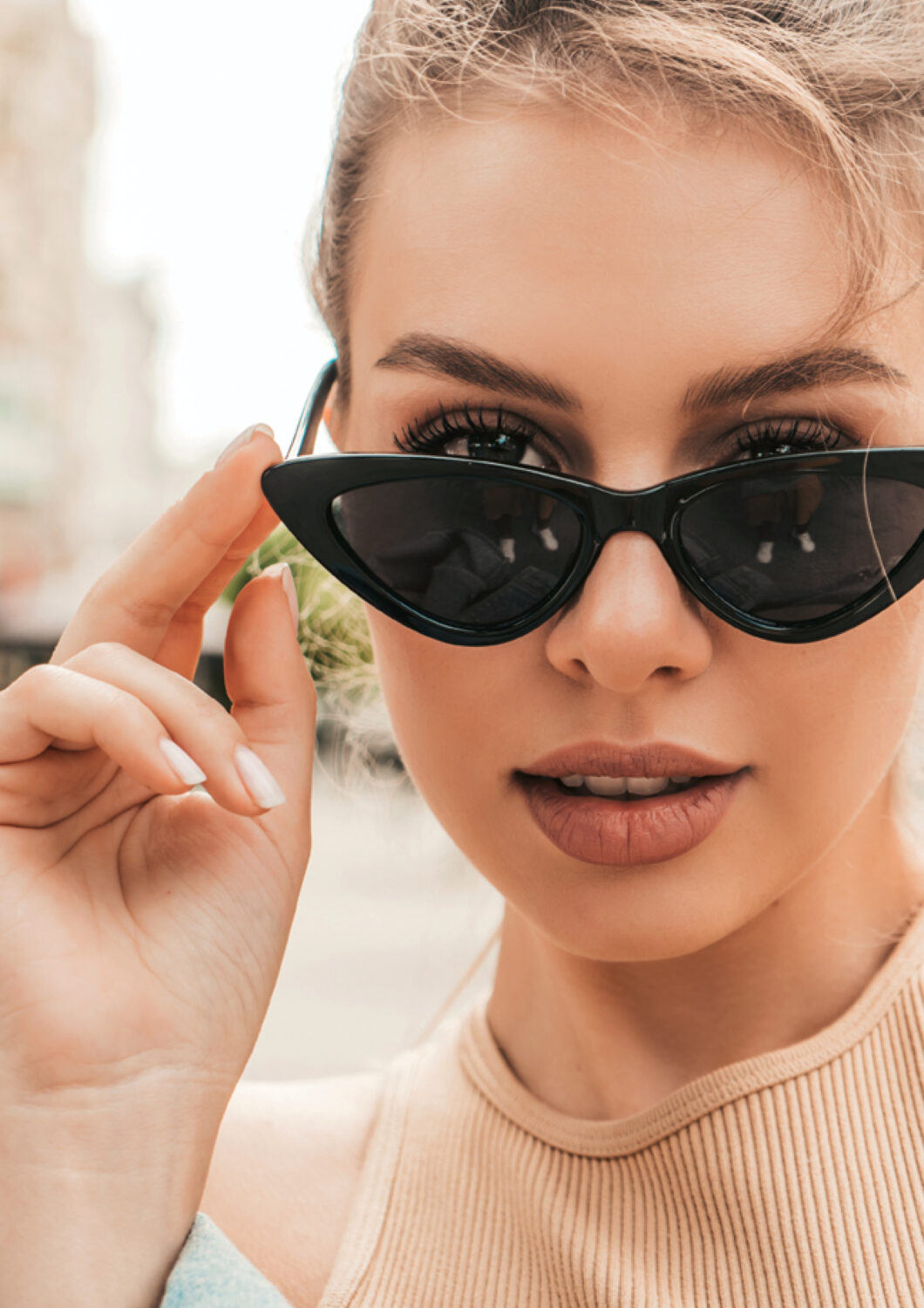Blonde woman wearing black Revex sunglasses in cat-eye model looks over the frame in an urban setting.