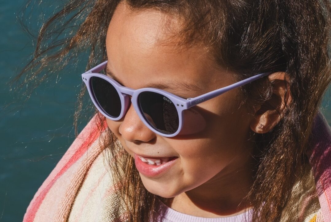 Smiling girl with purple children's sunglasses by Okky and beach towel by the water, ideal for wholesale.