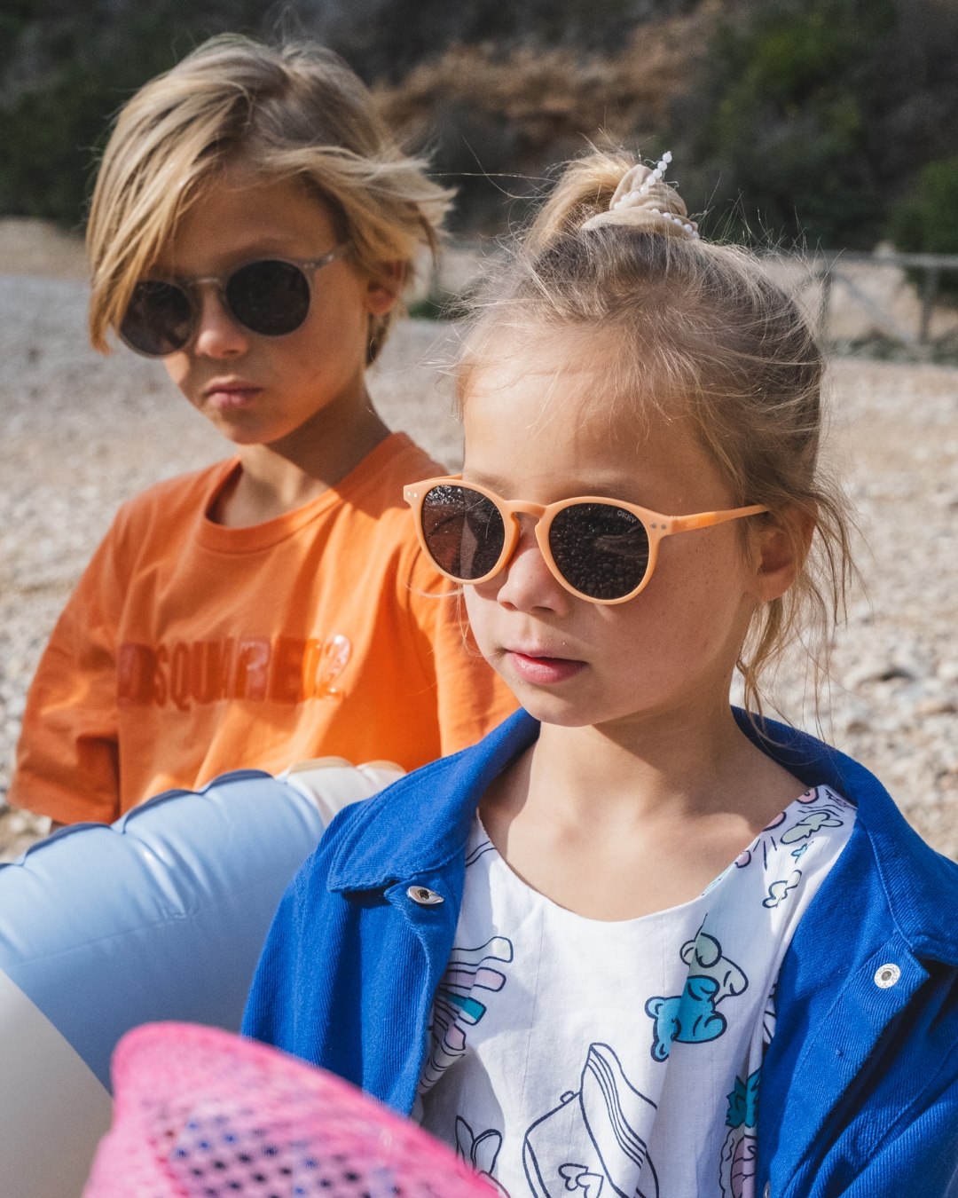 A blonde boy and girl wear trendy children's sunglasses by the brand Okky on a pebble beach during a sunny day.