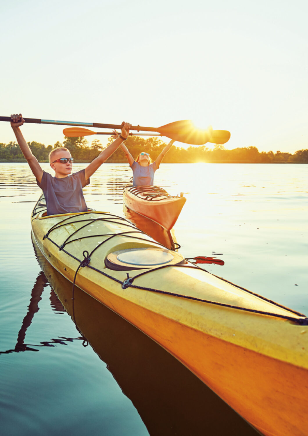 Two people kayaking on a calm lake at sunset, the boy in front wearing sports glasses.
