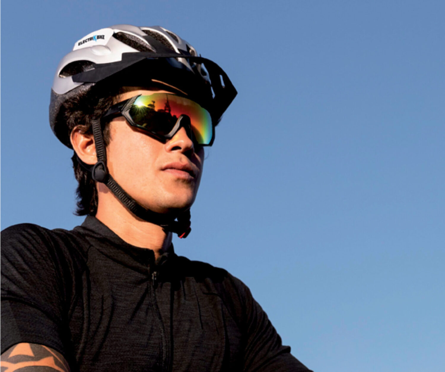Man on bike with Revex sports sunglasses, bike helmet and arm tattoos against a clear blue sky.