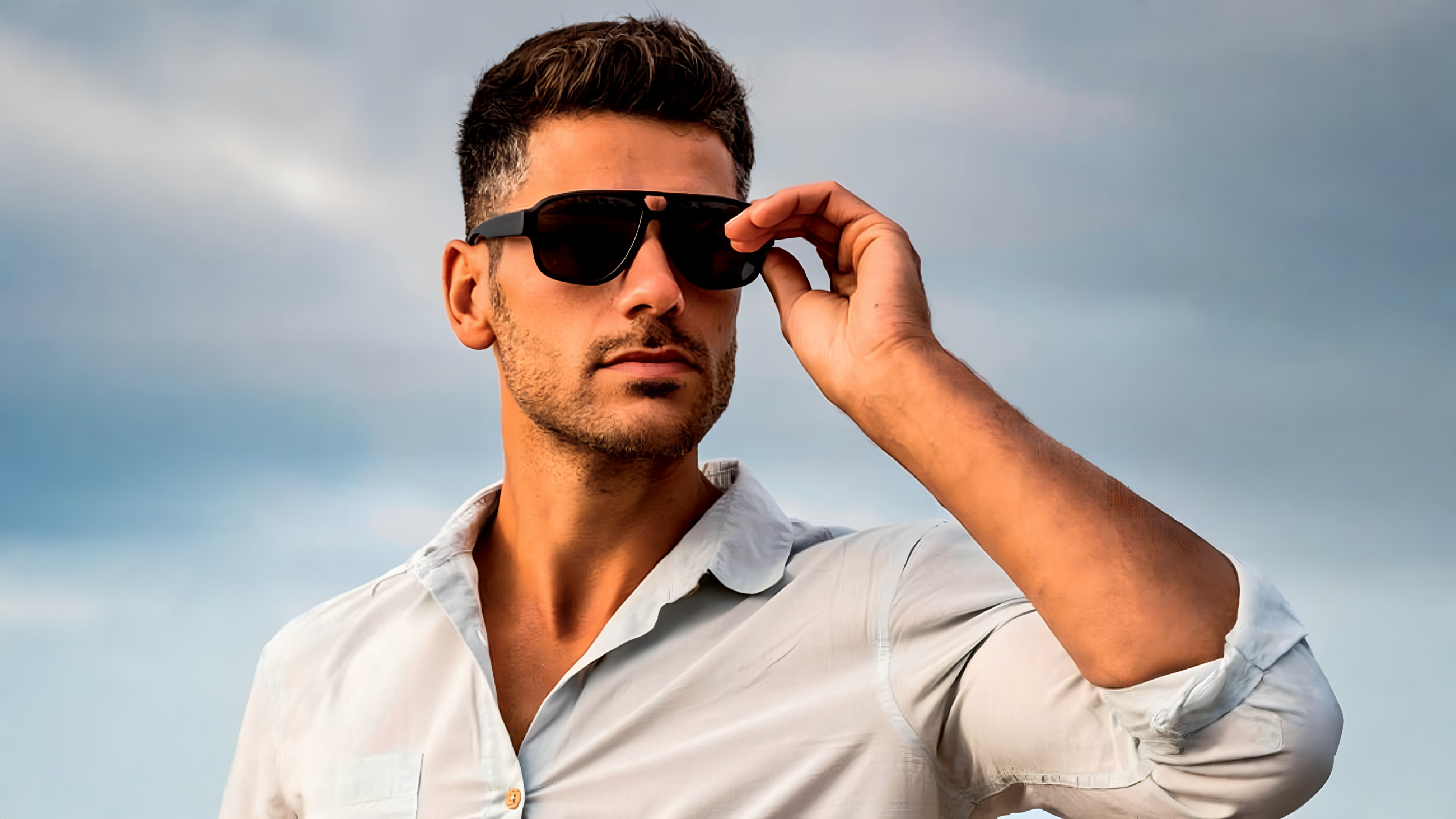 A man with stubble matches his black sunglasses against an overcast blue sky.