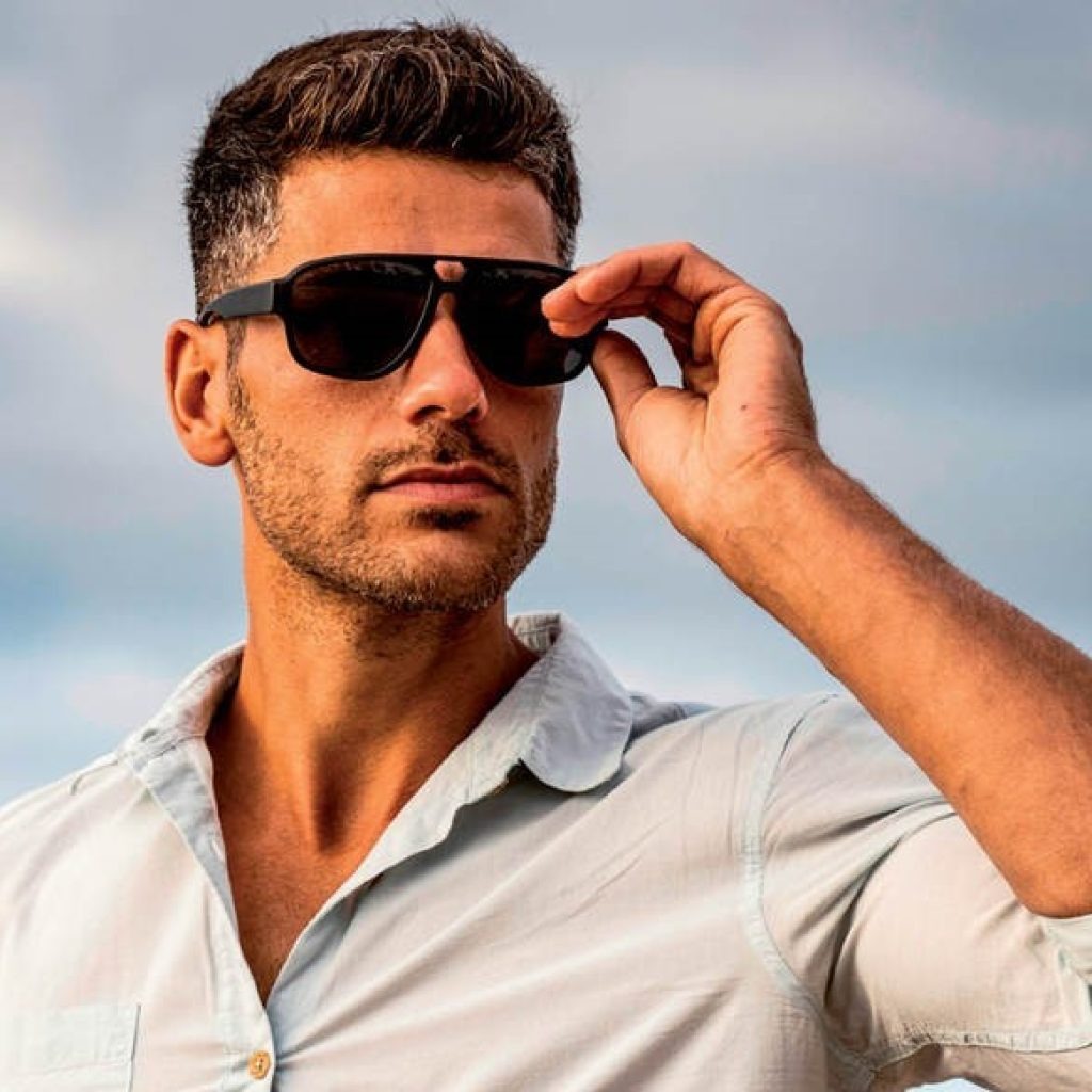 A stylish man wearing a light blue shirt straightening his black sunglasses against a clear sky.