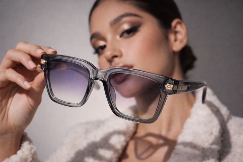 Una mujer sostiene unas gafas de sol cuadradas grises con montura transparente y cristales degradados para un retrato.