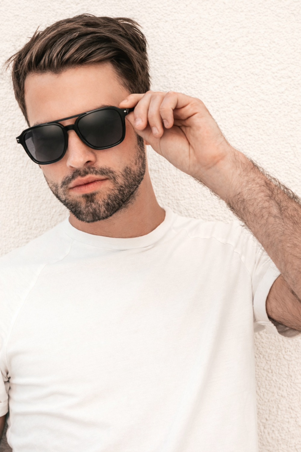Hombre elegante con gafas de sol negras y camiseta blanca contra una pared, foco en las gafas de sol masculinas.